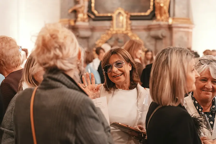Gäste im Gespräch vor einem goldverzierten Seitenaltar, eine Person hebt die Hand