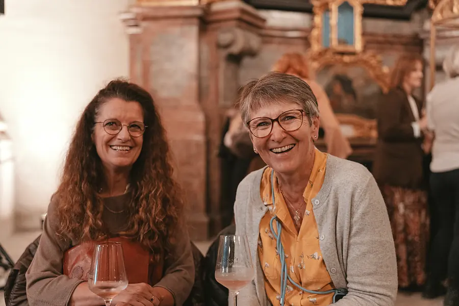 Zwei Gäste mit Weingläsern lachen am Tisch vor einem barocken Seitenaltar